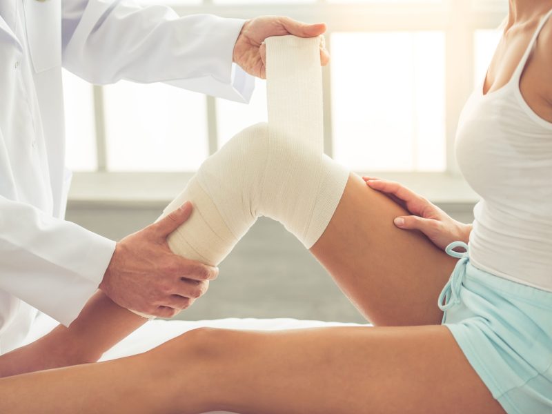 Cropped image of handsome doctor bandaging woman's injured knee while working in his office